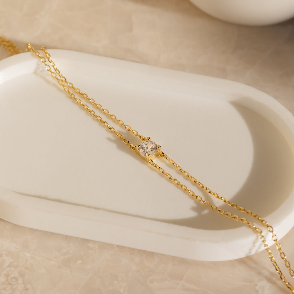 The Duo Chain Diamond Bracelet features a single clear gemstone on a gold double chain, elegantly displayed on a white tray—perfect as an anniversary gift.