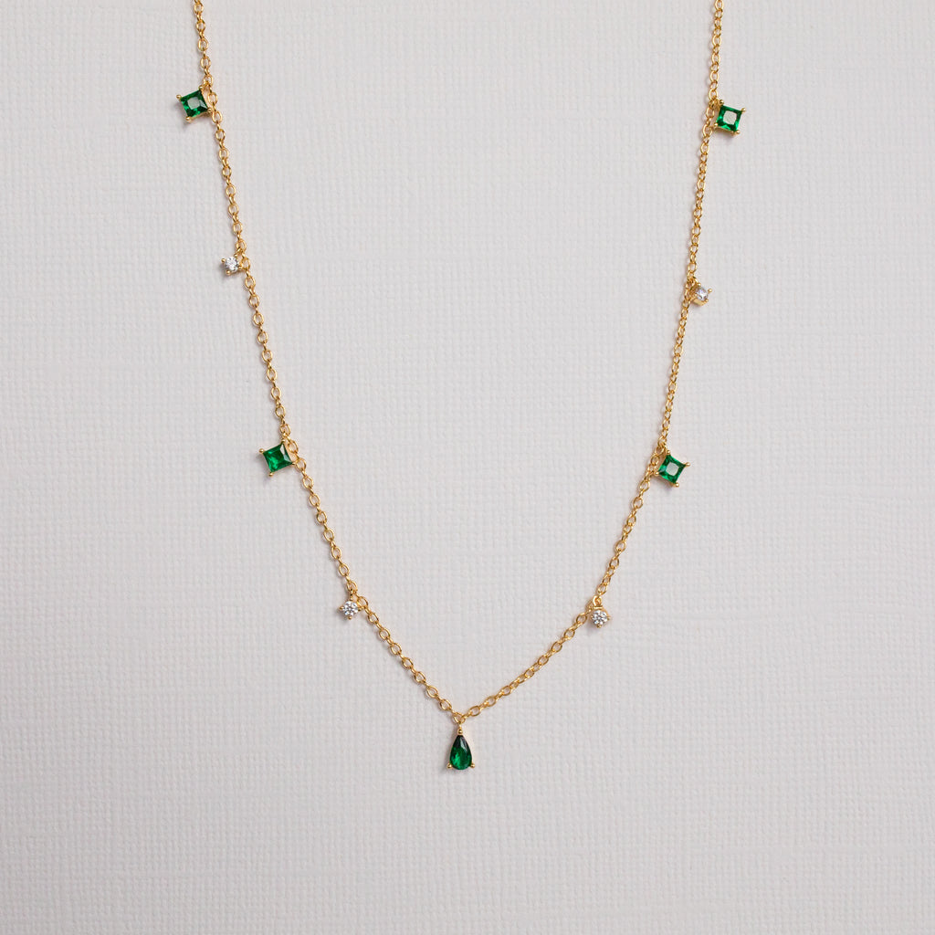 The Larissa Emerald Station Necklace features small green and clear gemstone charms spaced along a gold chain, offering a dainty, vintage-inspired look. Shown on a white background.