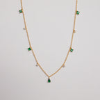 The Larissa Emerald Station Necklace features small green and clear gemstone charms spaced along a gold chain, offering a dainty, vintage-inspired look. Shown on a white background.