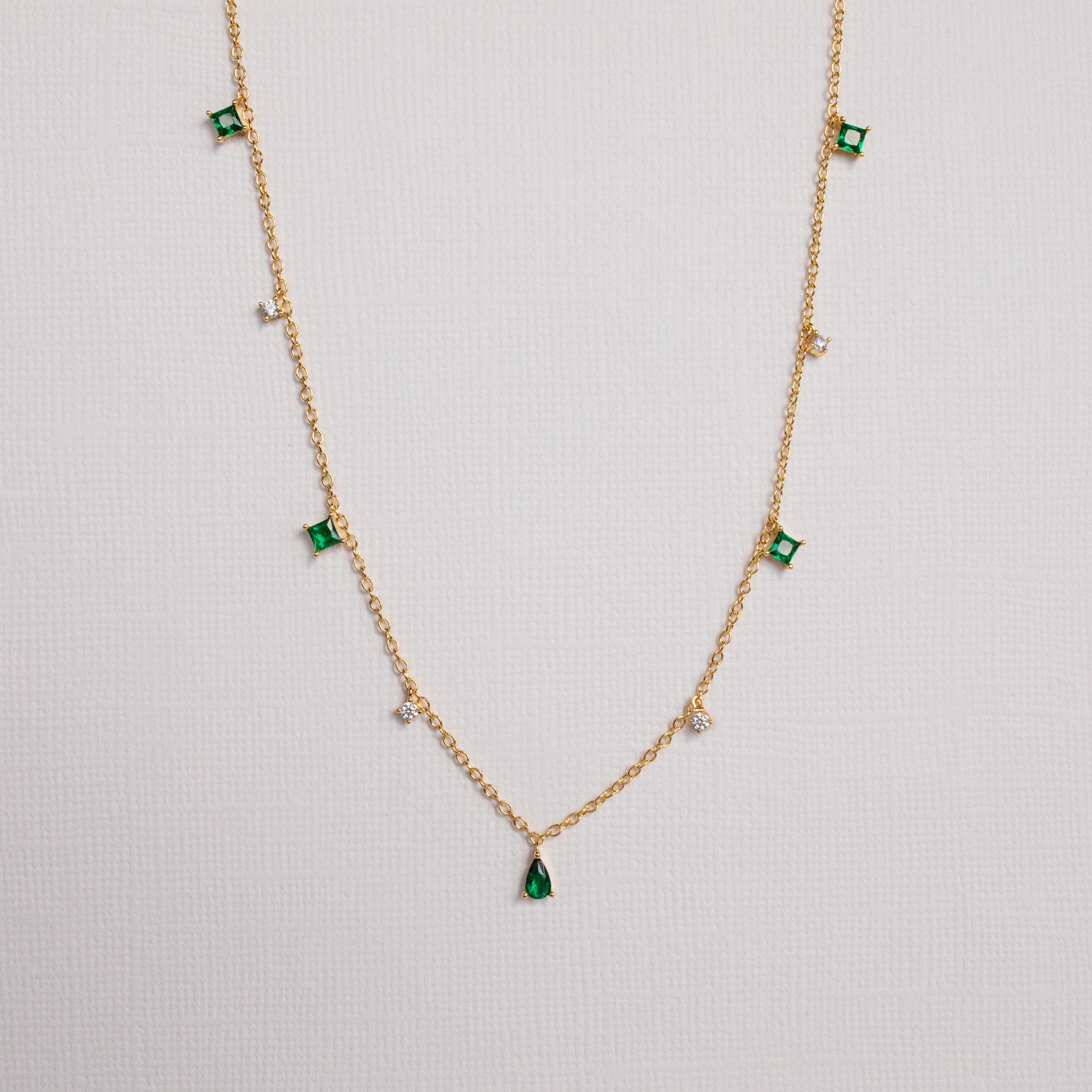 The Larissa Emerald Station Necklace features small green and clear gemstone charms spaced along a gold chain, offering a dainty, vintage-inspired look. Shown on a white background.