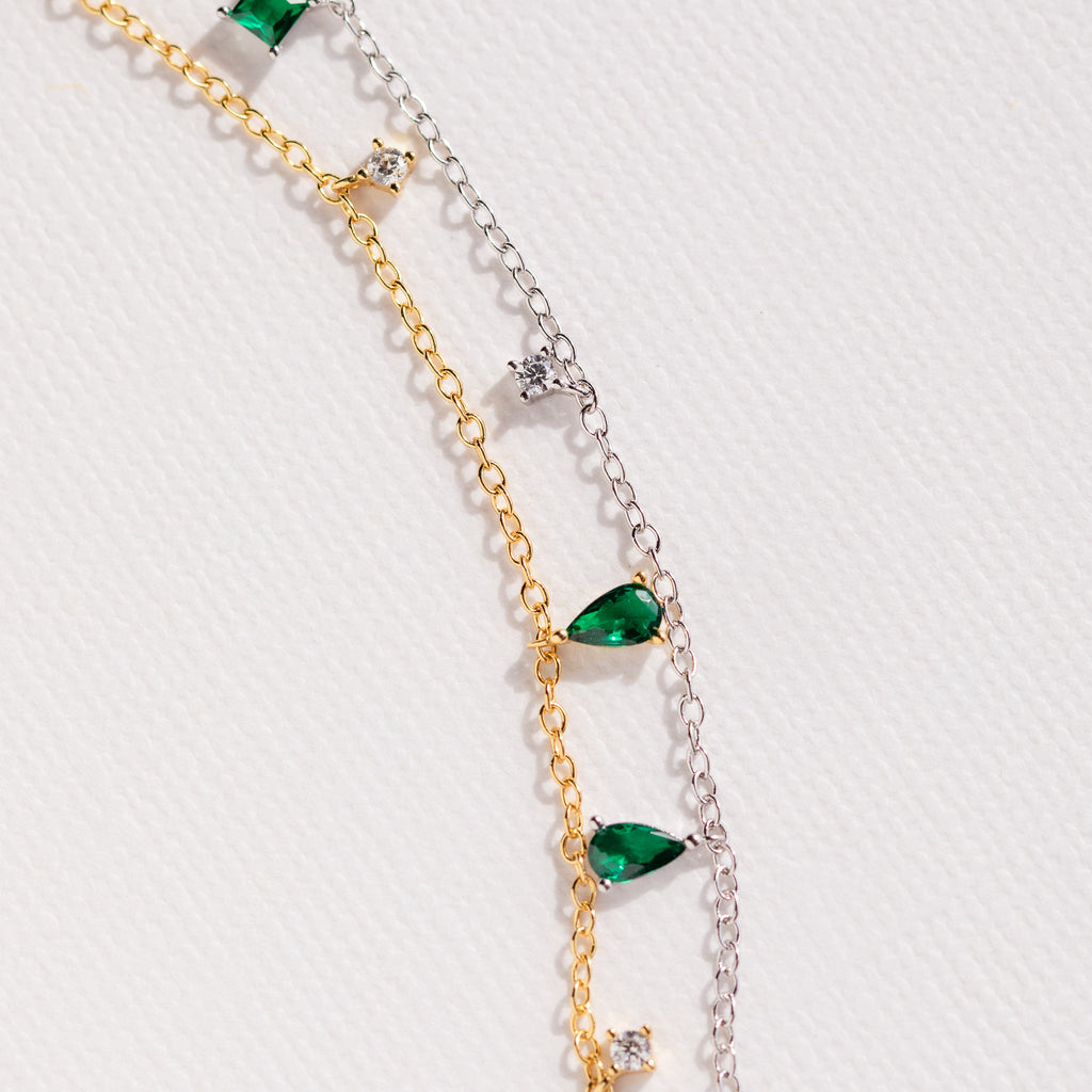 The Larissa Emerald Station Necklace features two delicate chains—one gold, one silver—adorned with green teardrop and clear round gemstones for a dainty, vintage-inspired look, perfect for layering or wearing solo.