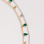 The Larissa Emerald Station Necklace features two delicate chains—one gold, one silver—adorned with green teardrop and clear round gemstones for a dainty, vintage-inspired look, perfect for layering or wearing solo.