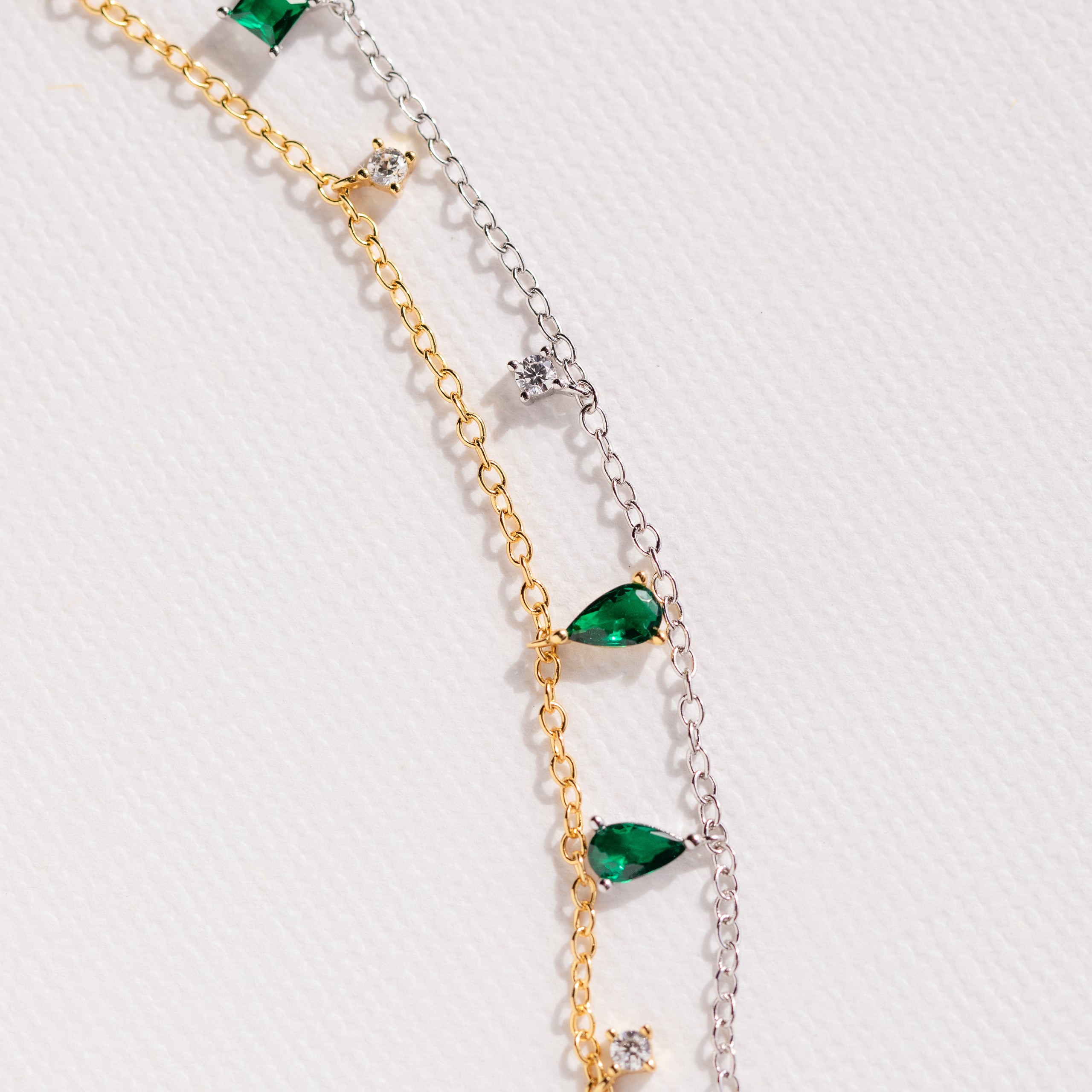 The Larissa Emerald Station Necklace features two delicate chains—one gold, one silver—adorned with green teardrop and clear round gemstones for a dainty, vintage-inspired look, perfect for layering or wearing solo.