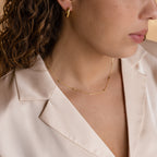 A woman is shown from neck to shoulder, wearing a beige blouse, gold hoop earrings, and the Bar Station Necklace with delicate chains.