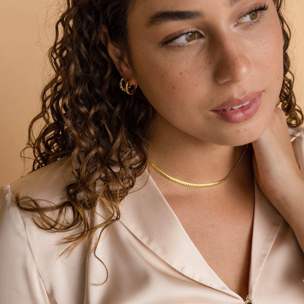 Woman with curly hair wearing gold jewelry, featuring the Thick Snake Chain Necklace, and a satin blouse, looking thoughtfully to the side.
