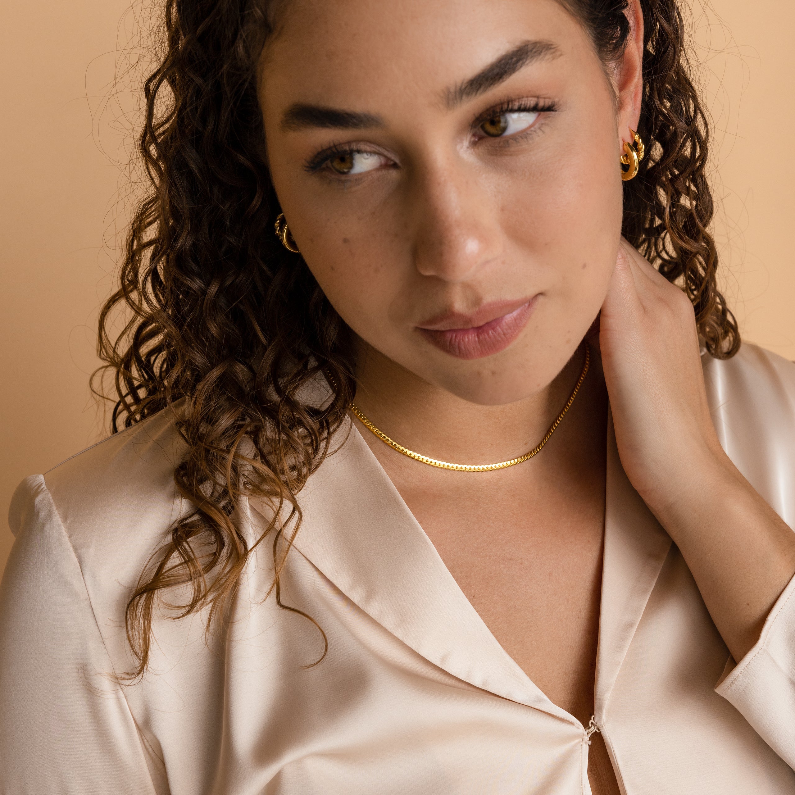 Woman with curly hair wears a Thick Snake Chain Necklace and gold jewelry with a cream blouse, touching her neck while looking to the side.