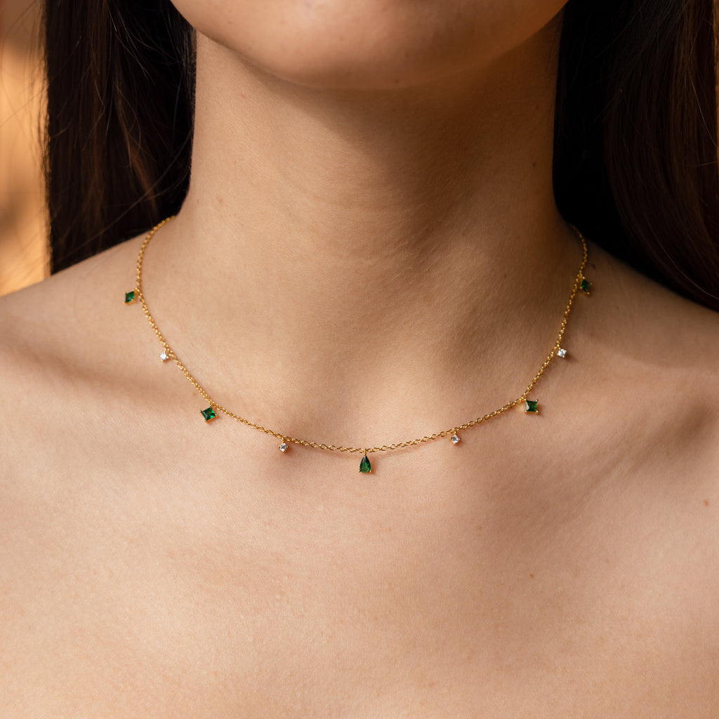 A woman wears the Larissa Emerald Station Necklace—a delicate gold chain featuring dainty green and clear gemstone charms in a vintage-inspired design.