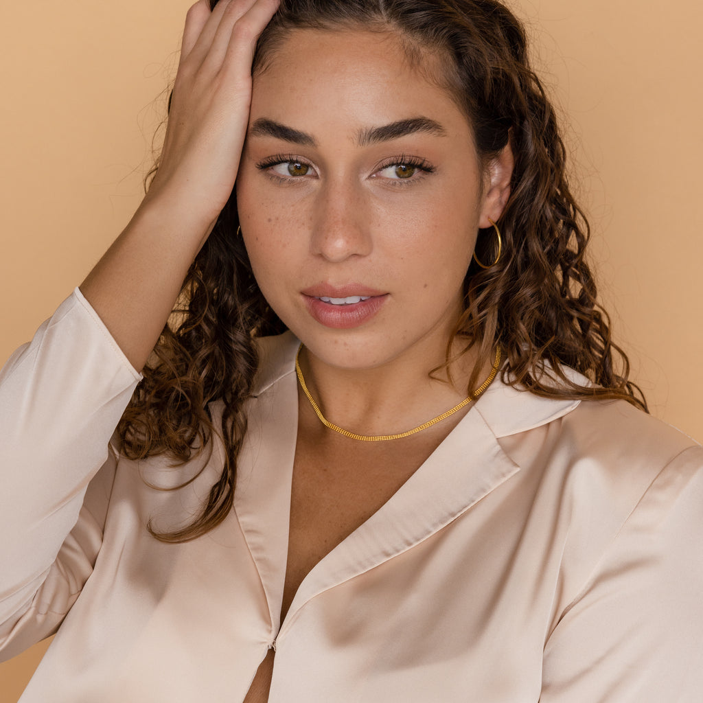 Woman with curly brown hair in a cream satin blouse, gold hoop earrings, and the Tri-Stacked Beaded Necklace touches her head.