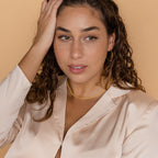 Woman with curly brown hair in a cream satin blouse, gold hoop earrings, and the Tri-Stacked Beaded Necklace touches her head.