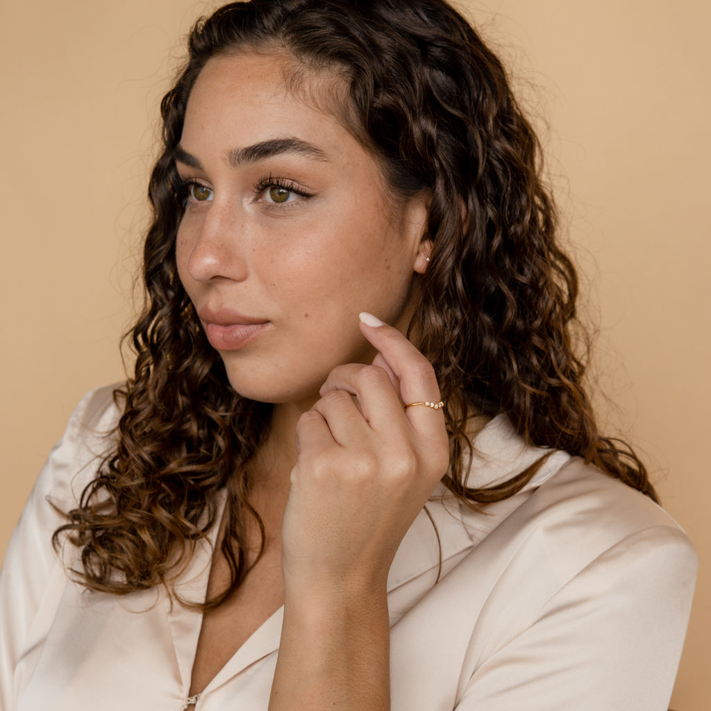 Woman with curly brown hair in a cream blouse looks to the side, gently touching her face with a hand adorned by the Layla Opal Diamond Ring.