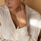 A woman in a white pleated blouse touches her earring, elegantly showcasing our Duo Chain Diamond Bracelet—a minimalist piece that's the perfect anniversary gift.