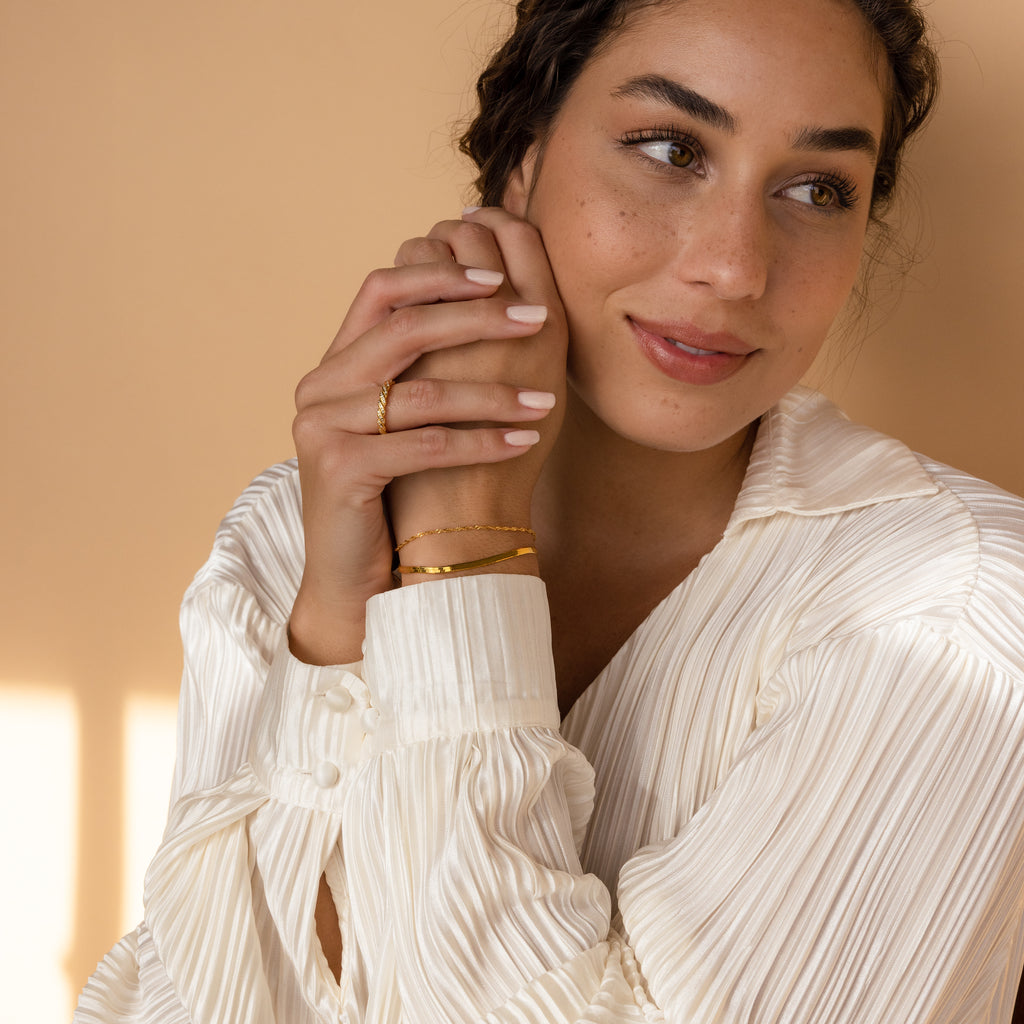 A woman in a white pleated blouse, smiling softly, wears gold jewelry, including the Singapore Twist Bracelet, with her hands gently clasped.