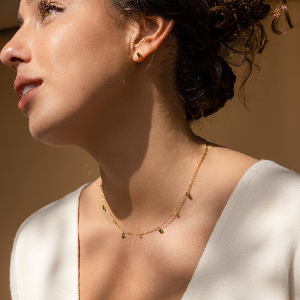 A woman wears the Larissa Emerald Station Necklace with green stones and matching stud earrings, photographed in soft natural light.