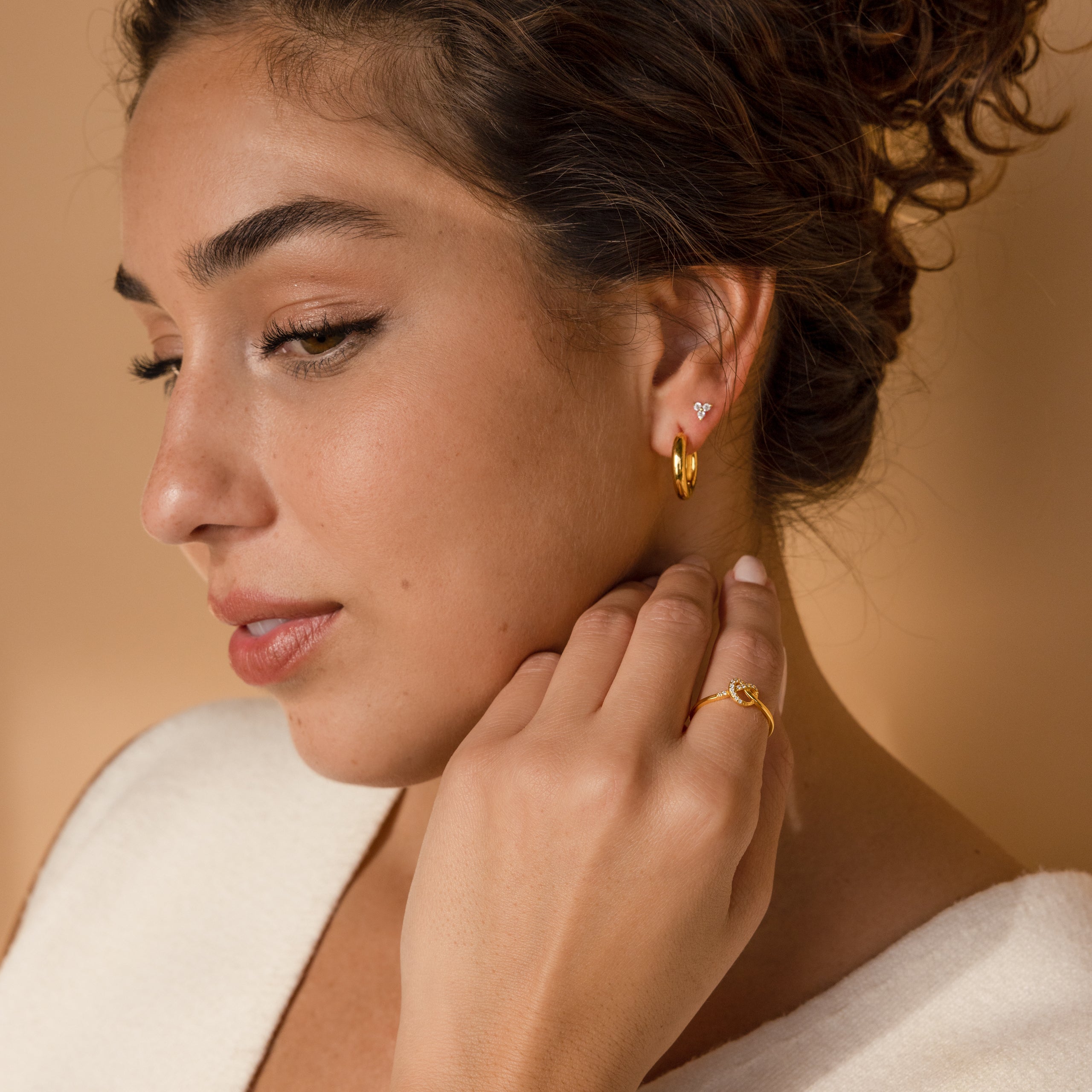 Woman with curly hair wearing gold hoop earrings and a Pave Heart Knot Ring, looking contemplative against a beige background—a perfect promise ring and romantic gift.