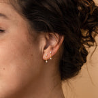 Close-up of a woman's ear with two gold earrings—a hoop and the Opal Drop Studs—against a neutral background. A dainty chain enhances the opal gemstone, adding an elegant touch.