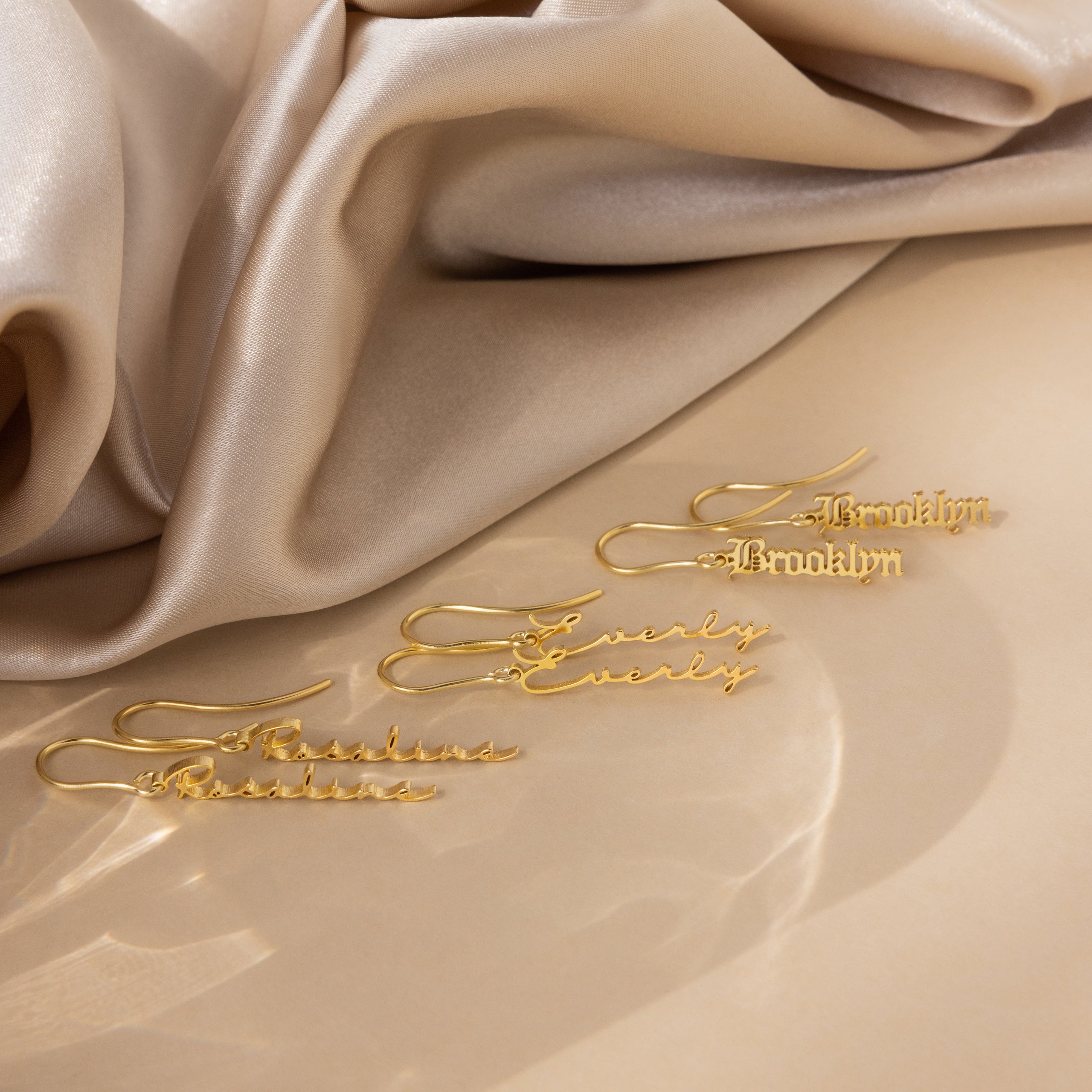 Three Old English Dangling Name Earrings reading Brooklyn and Everly rest on beige satin—perfect as custom, handmade drop earrings or a personalized gift for someone special.