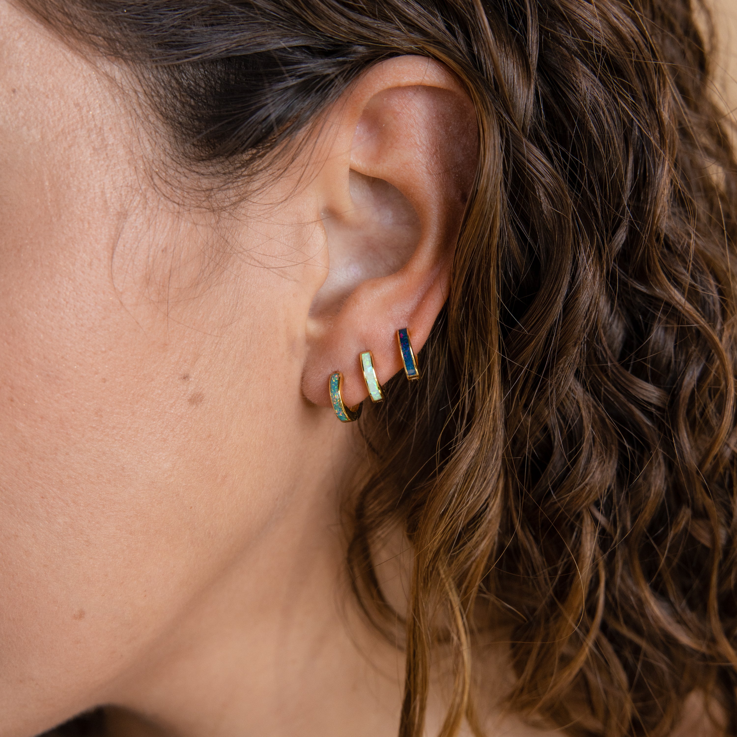 A close-up of a woman's ear wearing the Opal Inlay Huggies Set—three colorful hoop earrings with opal accents—complemented by her curly brown hair.