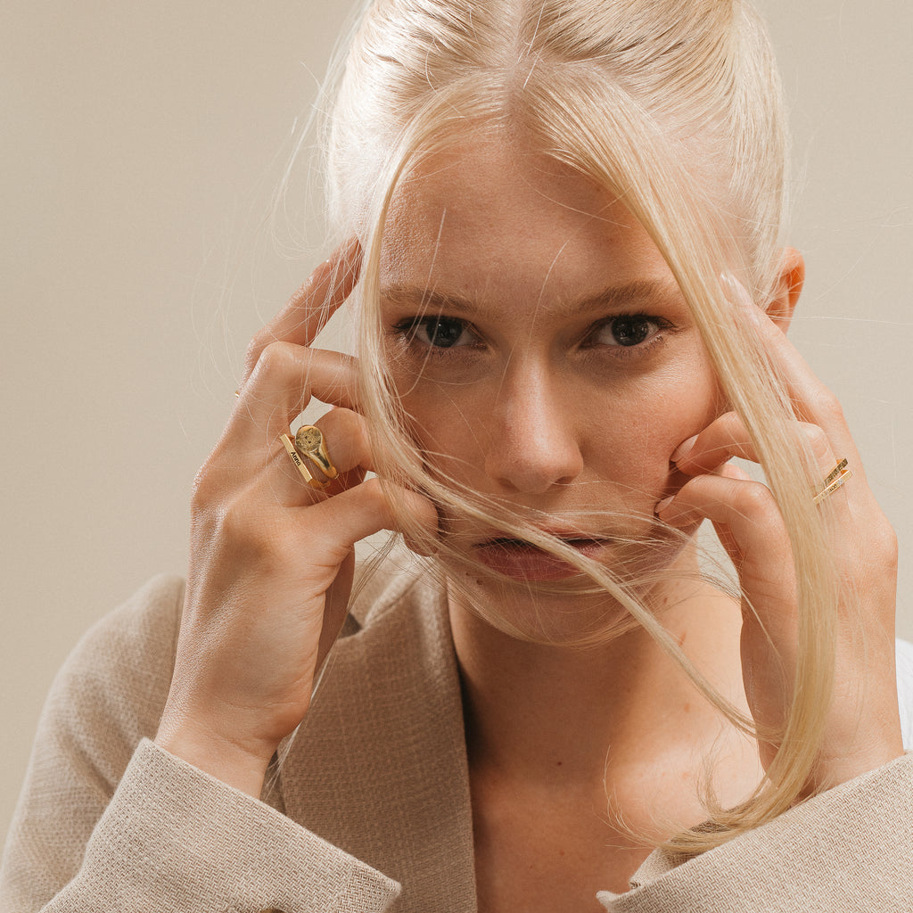 A blonde woman with loose hair touches her face, wearing gold rings—including the Pet Signet Ring—and a beige blazer.