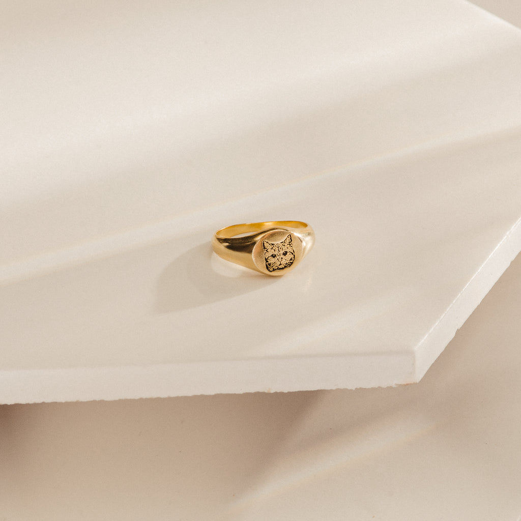 The Pet Signet Ring, engraved with a cat face, sits on a cream geometric surface—an adorable and personalized piece of pet jewelry.