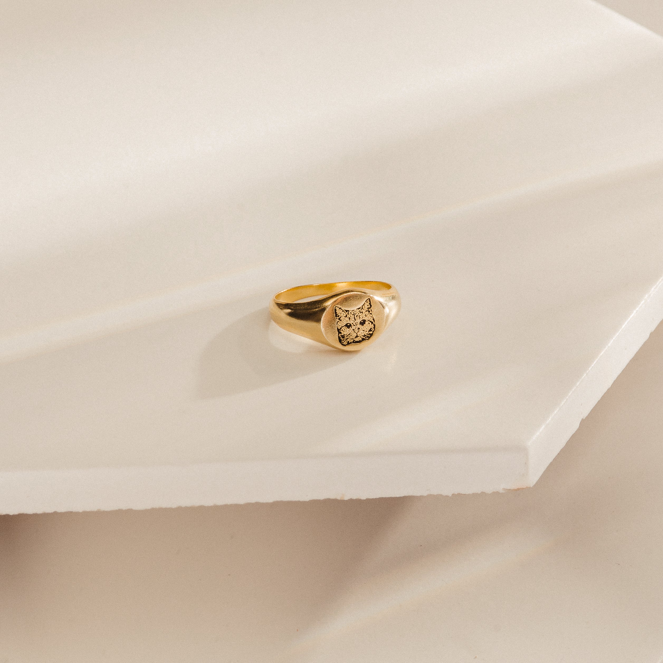 The Pet Signet Ring, engraved with a cat face, sits on a cream geometric surface—an adorable and personalized piece of pet jewelry.