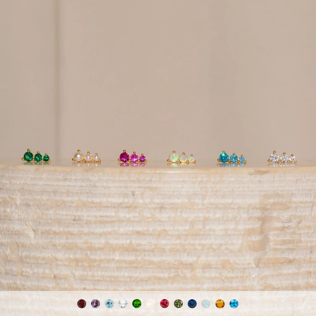 Six pairs of Triple Birthstone Studs are displayed on a light wooden surface with gem color icons below—making them a thoughtful gift for mothers and daughters.