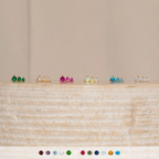 Six pairs of Triple Birthstone Studs are displayed on a light wooden surface with gem color icons below—making them a thoughtful gift for mothers and daughters.