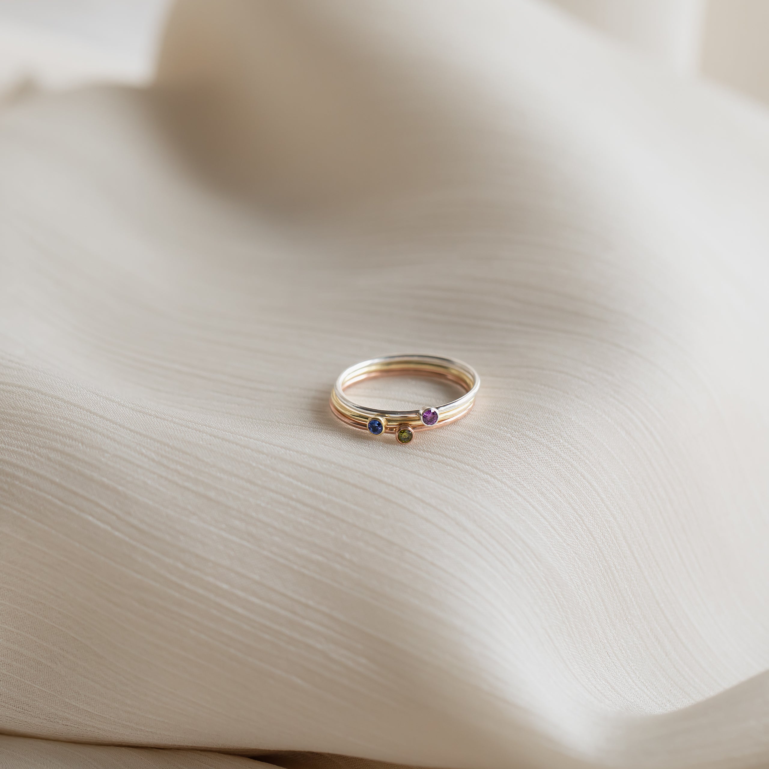 The Tiny Birthstone Ring sits on a white cloth, its delicate gemstone reflecting a subtle, colorful birthstone hue.