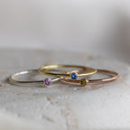 Three delicate Tiny Birthstone Rings showcase gemstone detailing: silver with a purple stone, gold with blue, and rose gold with green.