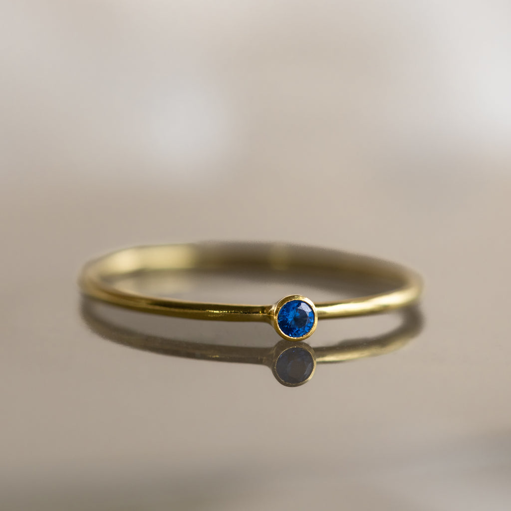 The Tiny Birthstone Ring is a thin gold ring with a small round blue gemstone and delicate gem detailing, shown on a reflective surface.