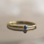 The Tiny Birthstone Ring is a thin gold ring with a small round blue gemstone and delicate gem detailing, shown on a reflective surface.