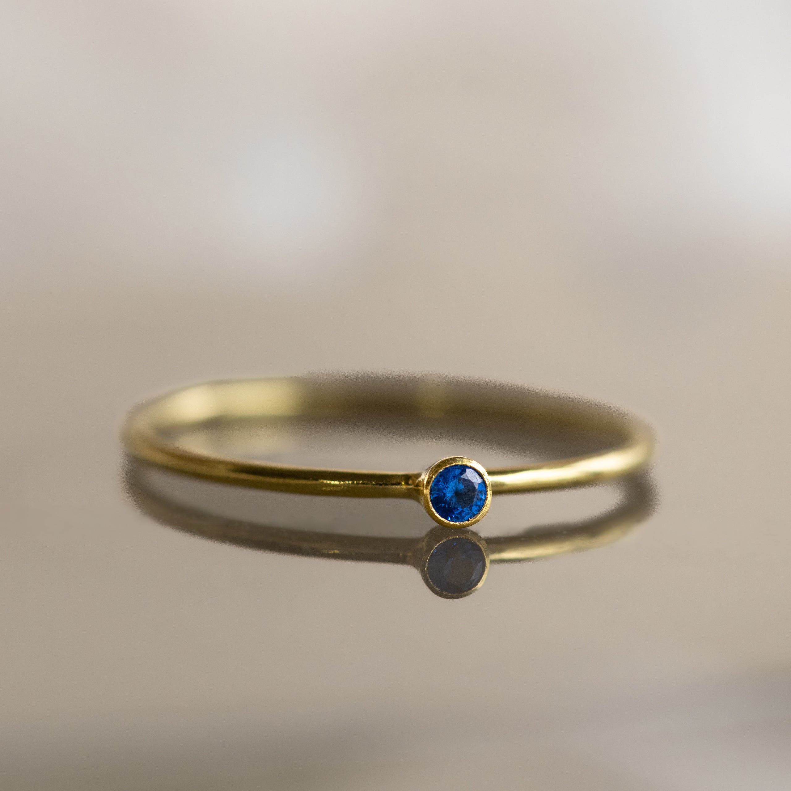 The Tiny Birthstone Ring is a thin gold ring with a small round blue gemstone and delicate gem detailing, shown on a reflective surface.
