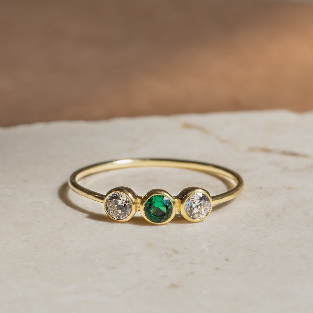 The Triple Birthstone Ring, featuring a central green stone flanked by two clear stones, rests beautifully on a light surface.