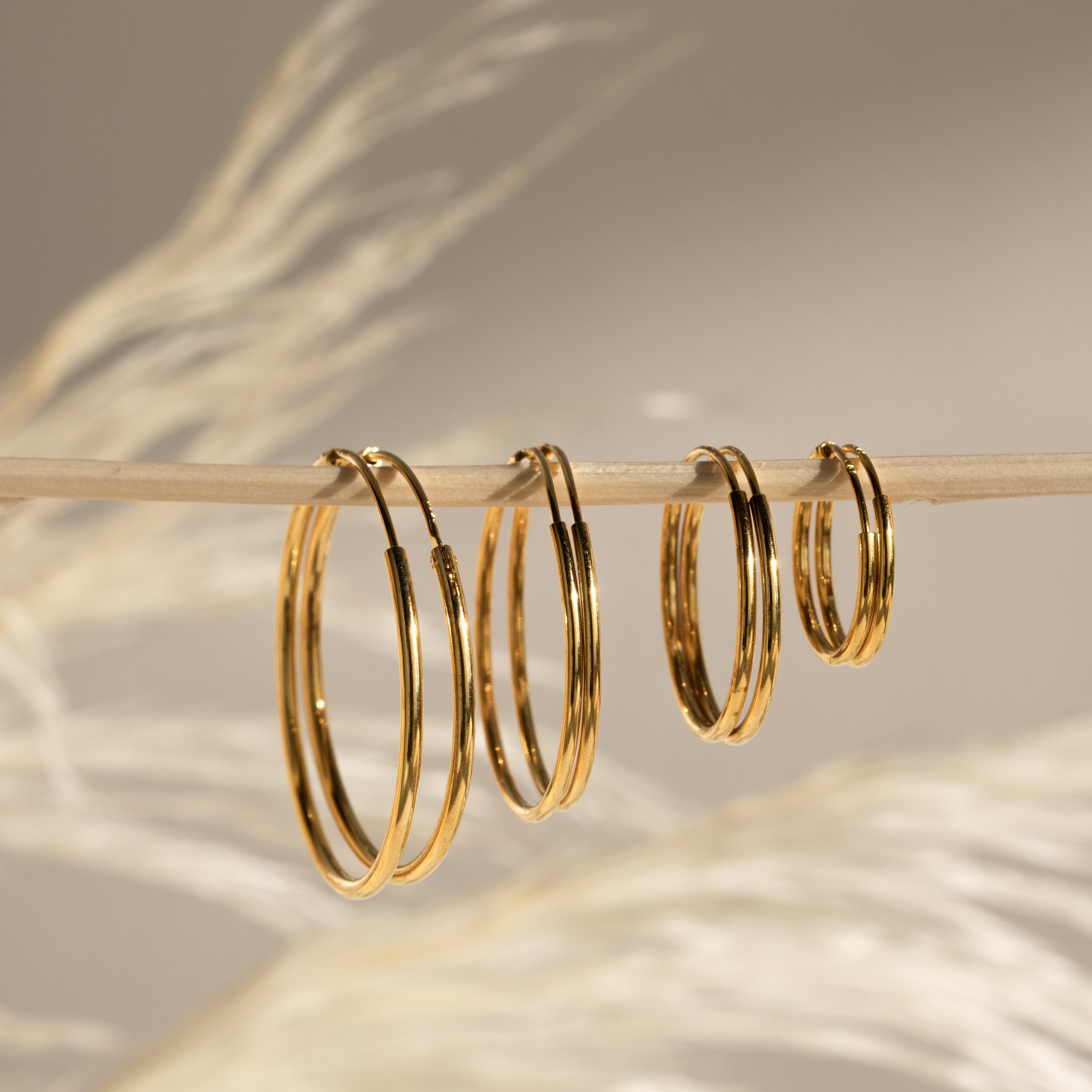 Five pairs of Endless Thin Hoops earrings in different sizes hang on a thin wooden stick against a blurred beige background—a perfect everyday accessory.