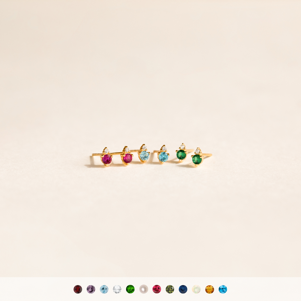 Five Raindrop Birthstone Studs featuring colorful gemstones are displayed on a light background, paired with dazzling diamond earrings and vibrant gems arranged below.