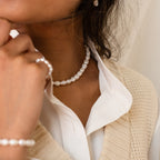 A woman touches her chin while wearing the Bold Pearl Necklace and a cream knit vest over a white shirt.