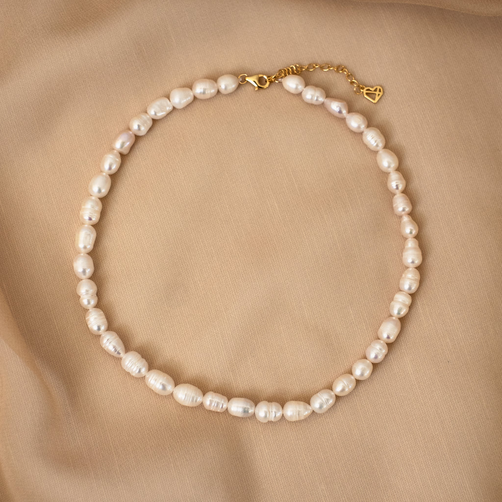 The Bold Pearl Necklace, featuring a gold clasp, is displayed on a beige fabric background—an elegant addition to your pearl jewelry collection.