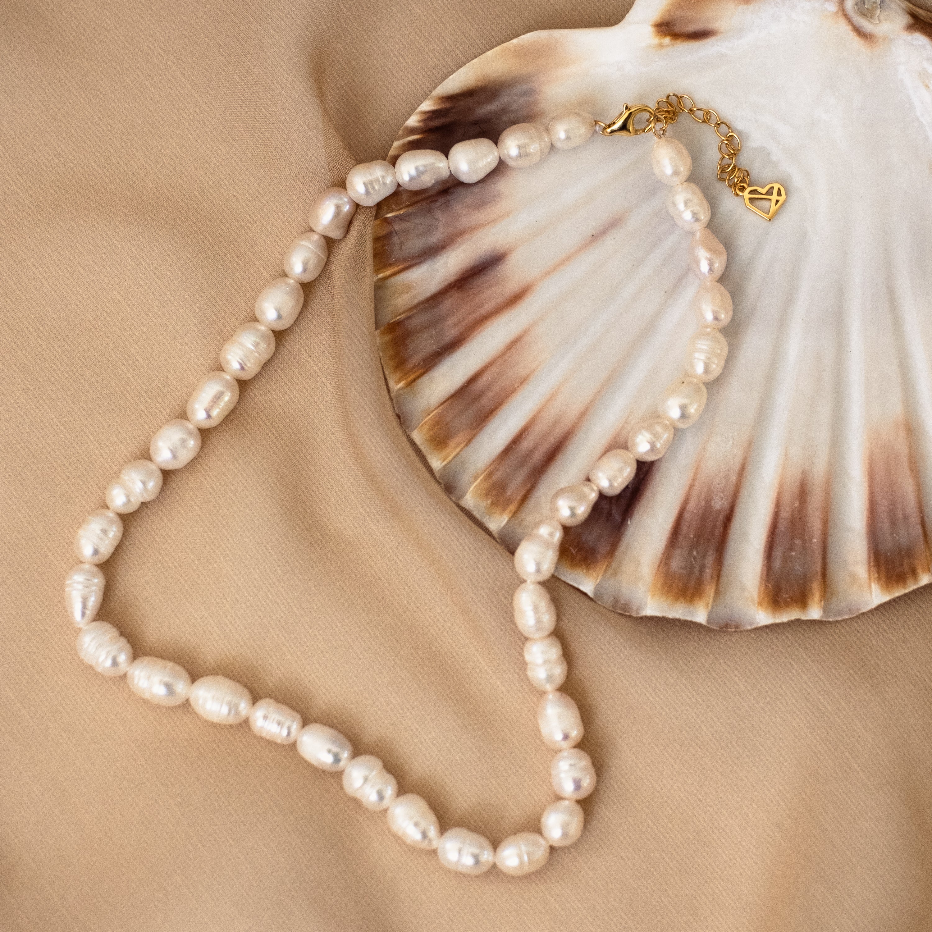 Bold Pearl Necklace made with irregular freshwater pearls, shown coiled beside a seashell on a beige fabric background—finished with a gold heart charm and clasp for an elegant touch.