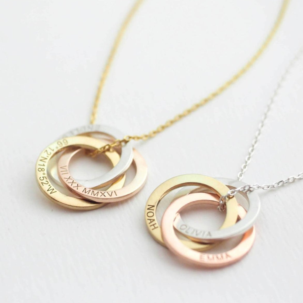 Two necklaces with interlocking circles in gold, silver, and rose gold, engraved with names, dates, and coordinates.