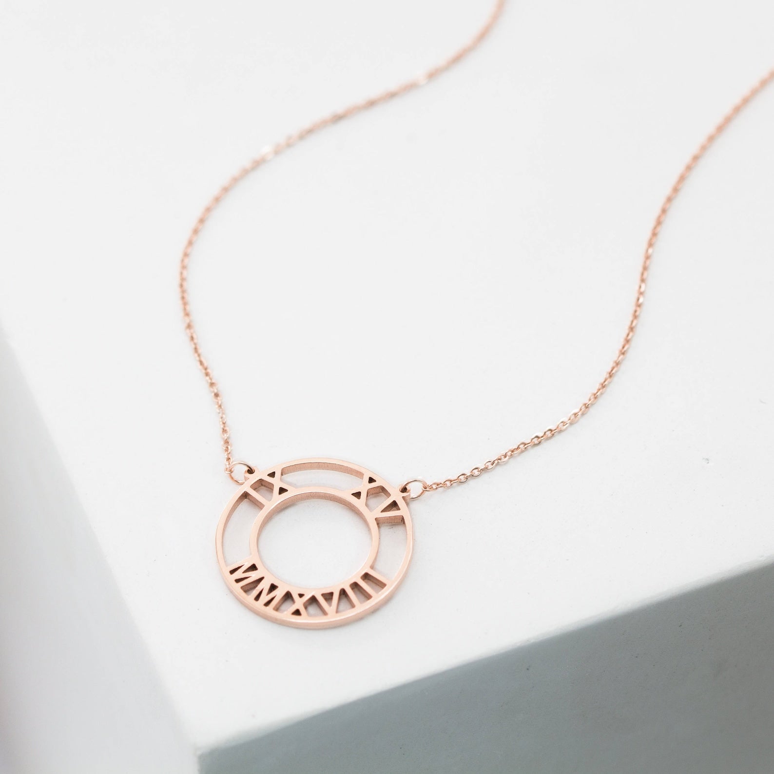 The Roman Numerals Necklace is a delicate rose gold piece with a circular geometric pendant engraved with Roman numerals, ideal as milestone jewelry and elegantly displayed on a white surface.