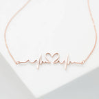 The Heartbeat Necklace in rose gold features a heartbeat line with a central heart, shown on a white background—a unique, personalized gift for someone special.