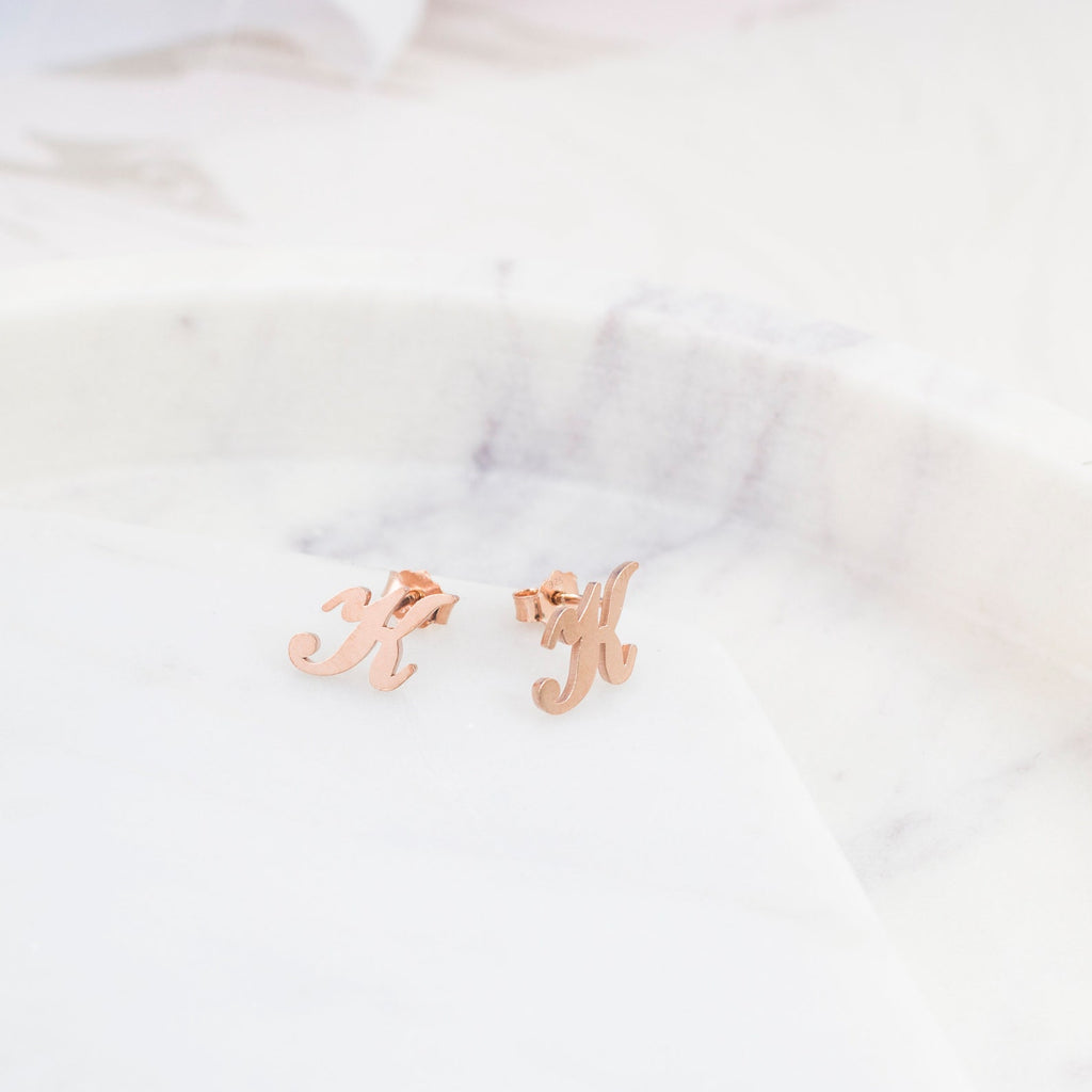Alex Initial Studs in rose gold, shaped as the letter H, rest elegantly on a white marble surface—perfect personalized earrings for any occasion.