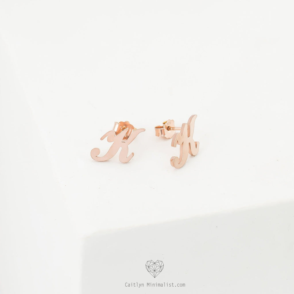 Alex Initial Studs in rose gold, shaped as the letter H, are displayed on a white surface—perfect dainty earrings for adding a personalized touch.