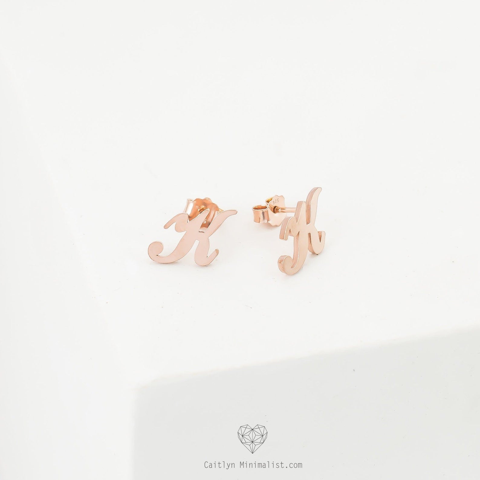Alex Initial Studs in rose gold, shaped as the letter H, are displayed on a white surface—perfect dainty earrings for adding a personalized touch.