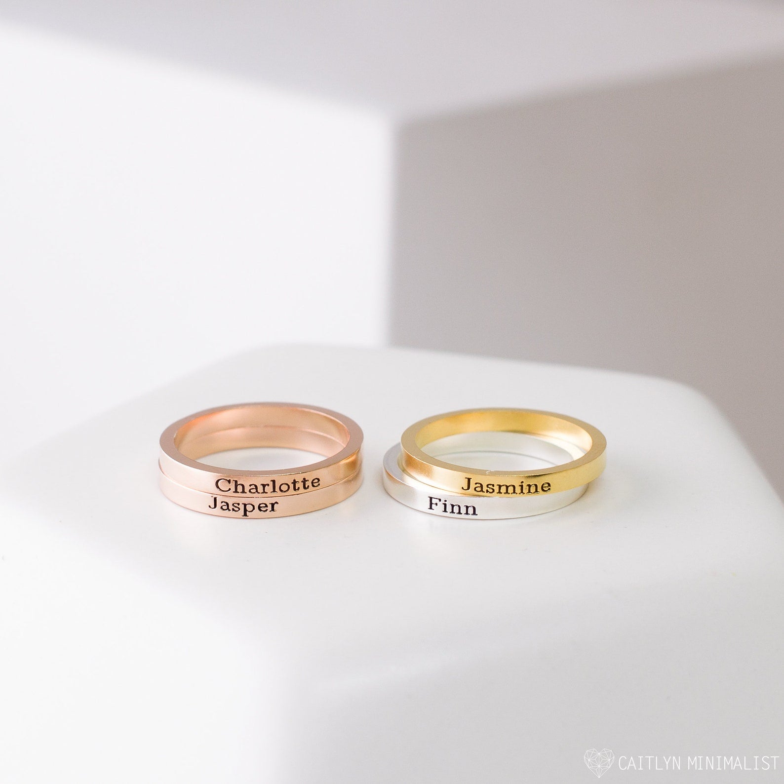 Two Stacking Name Band rings, custom engraved with names in rose gold and gold, are shown on a white surface—ideal as stackable rings for a personalized touch.
