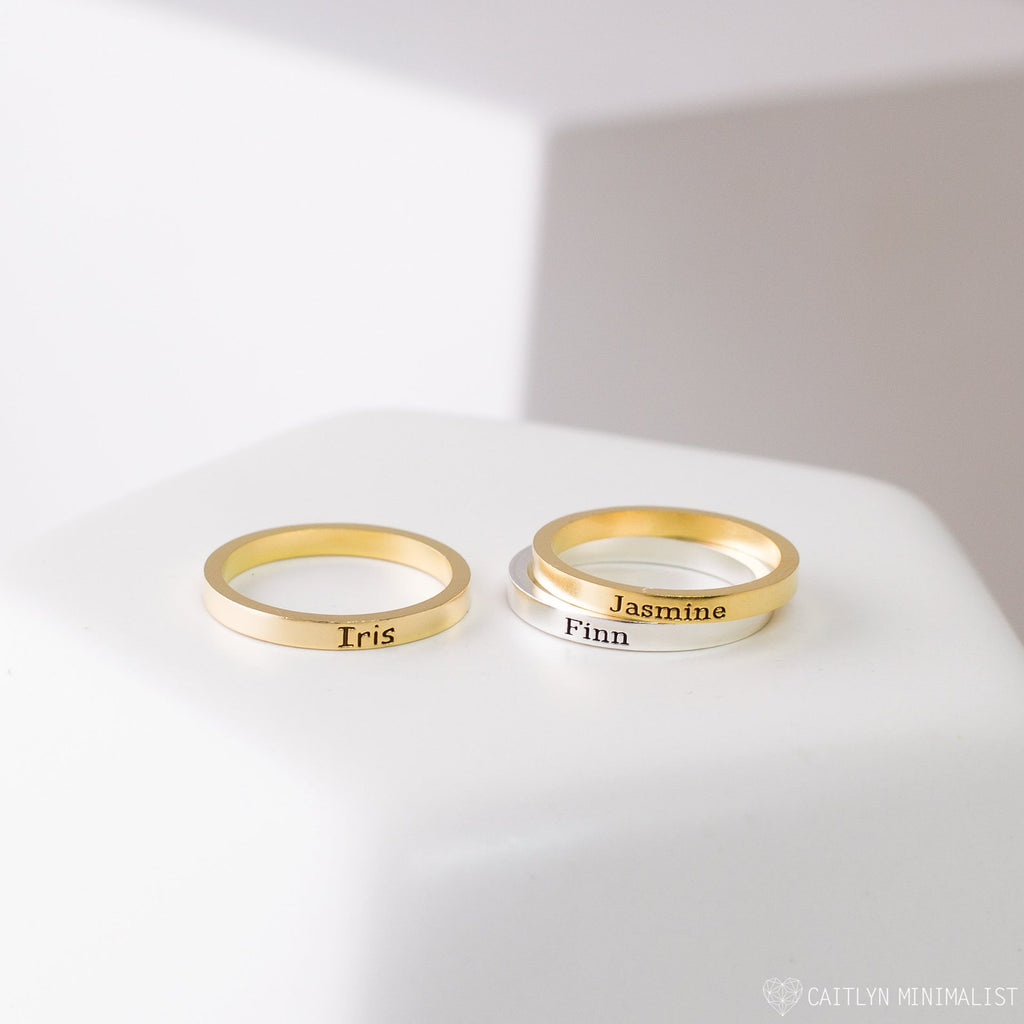 Three Stacking Name Bands—two gold and one silver—engraved with Tris, Jasmine, and Finn rest on a white surface, perfect for creating a personalized stackable ring set.