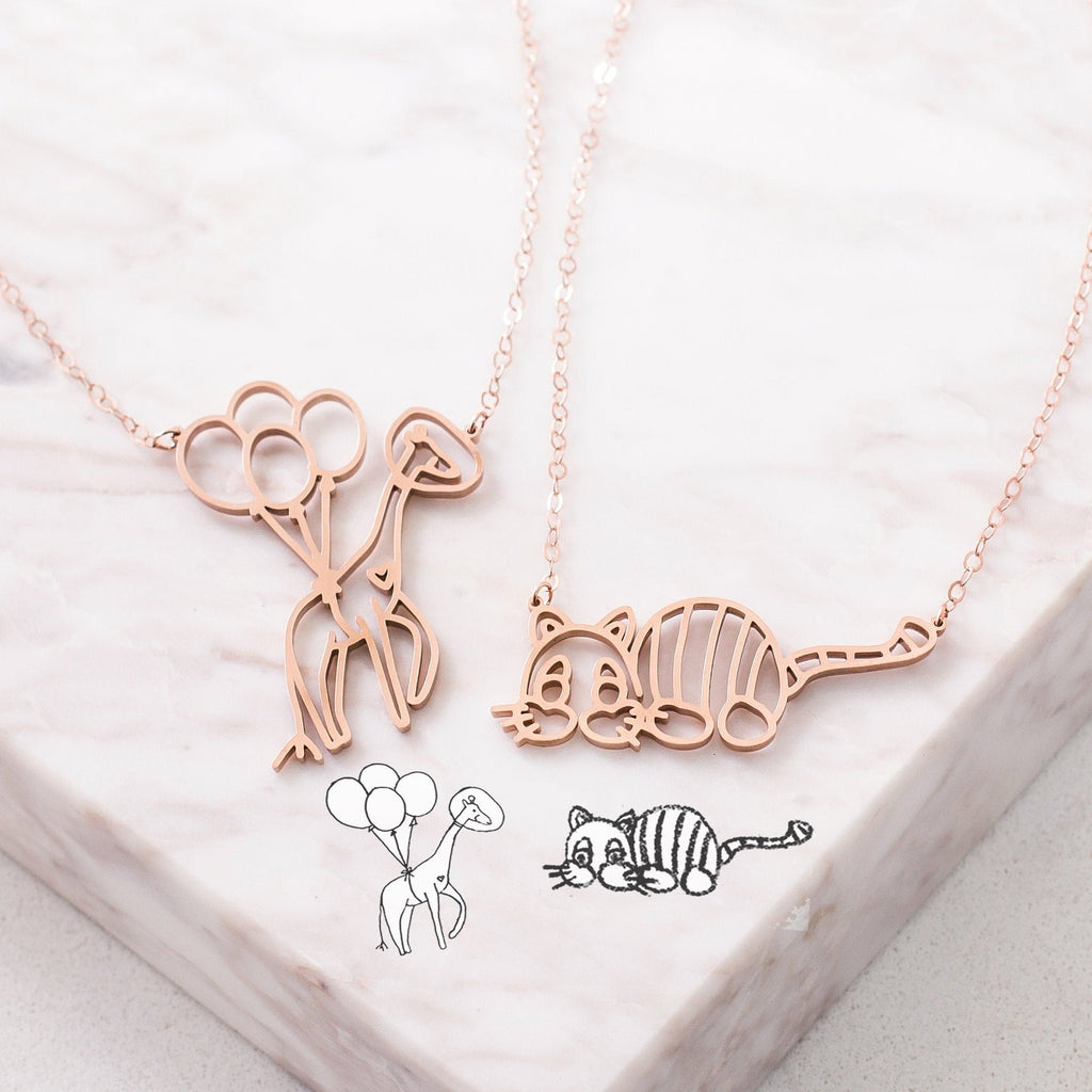 Two rose gold Kids Drawing Necklaces—one featuring a balloon dog, the other a cat—rest on marble. These unique pieces make perfect wearable art or personalized gifts for someone special.
