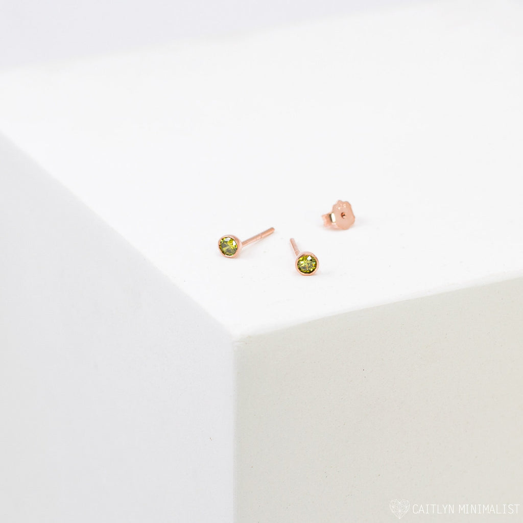 Birthstone Studs in rose gold with small green gemstones are displayed on a white geometric surface—ideal everyday earrings for a personalized style.