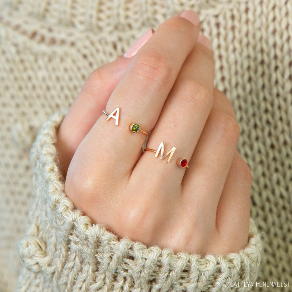 A hand with pink nails wears two Alex Initial Birthstone Rings, each featuring the letters A and M adorned with a small colored gem—stylish personalized wrap rings.