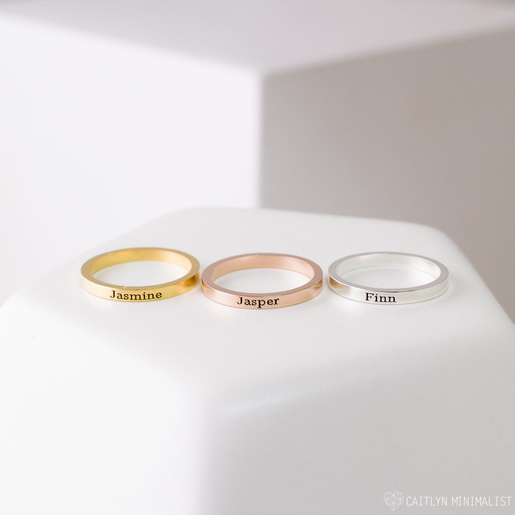 The Stacking Name Band features three custom engraved rings in gold, rose gold, and silver—shown with Jasmine, Jasper, and Finn—on a white surface. Ideal personalized jewelry for any meaningful occasion.
