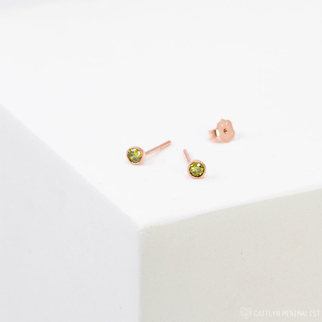 The Birthstone Studs are small, round gold earrings with green gemstones, shown on a white surface—ideal for everyday wear.